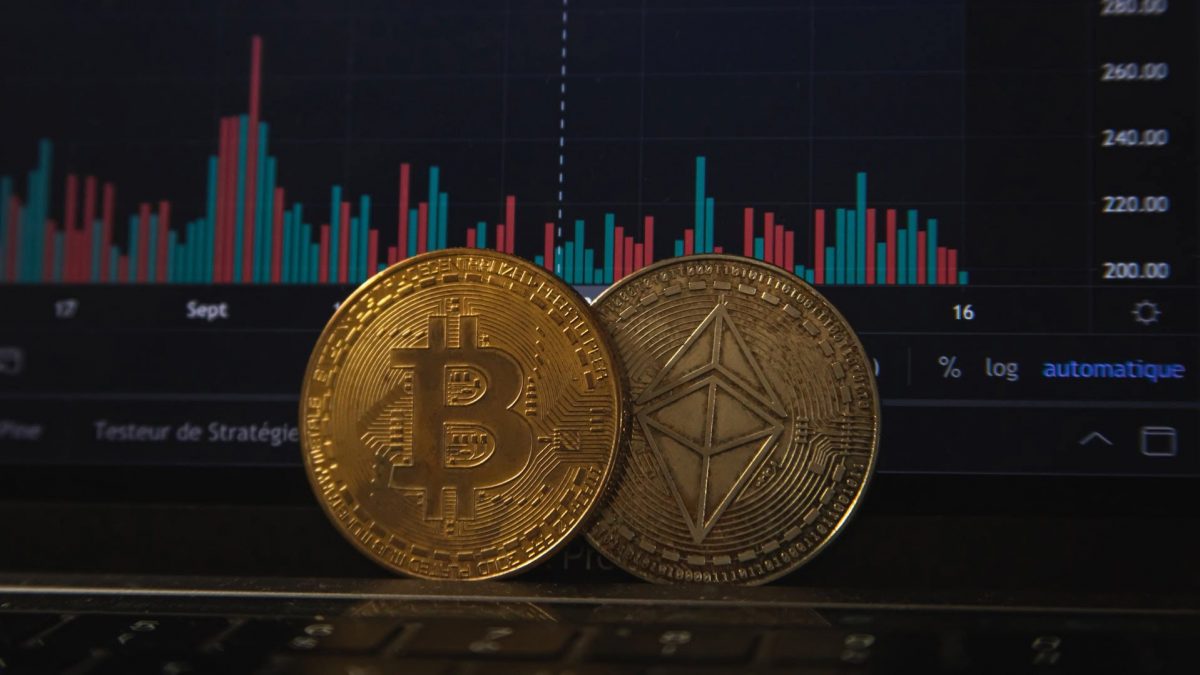 Physical coin representations of Bitcoin and Ethereum cryptocurrencies sit in front of a screen showing crypto value. 