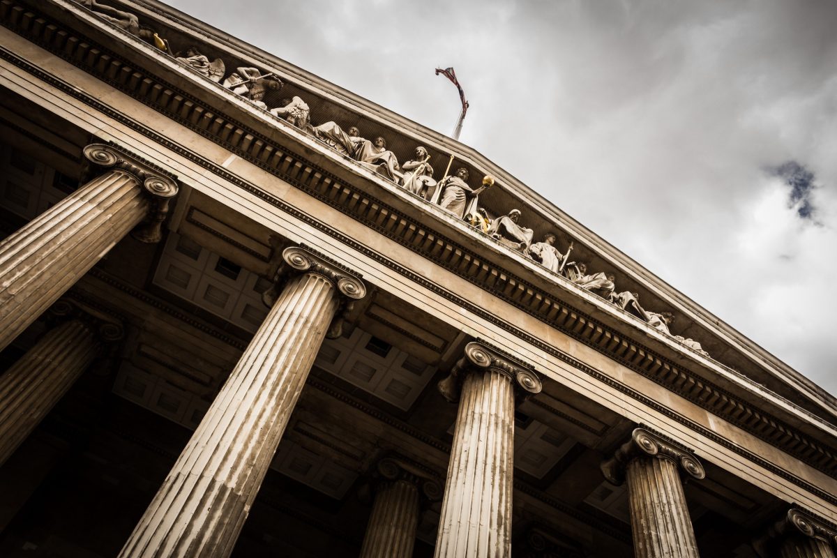 Stone columns rise up to support a wide roof of a building with cloudy skies in the background.