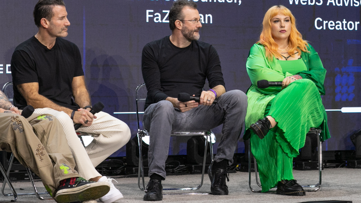 Una mujer con un vestido verde brillante y cabello rojizo sostiene un micrófono en un escenario junto a dos hombres con camisas negras.