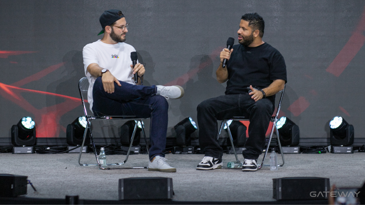 Two men sit on a stage talking to each other. One is wearing a white t-shirt, the other, a black t-shirt.