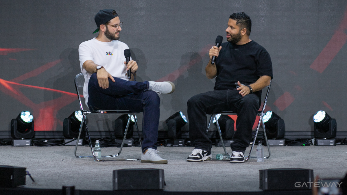 Two Men Sit On A Stage Talking To Each Other. One Is Wearing A White T-Shirt, The Other, A Black T-Shirt.