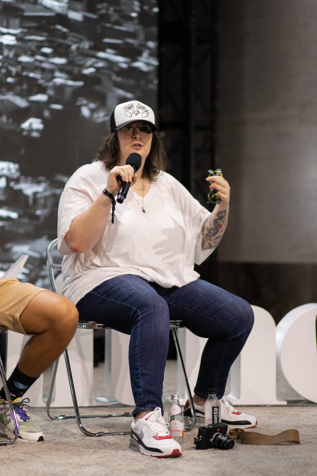 A woman in a baseball cap, white shirt, and jeans holds a microphone on a stage and gestures with her hand while speaking. 