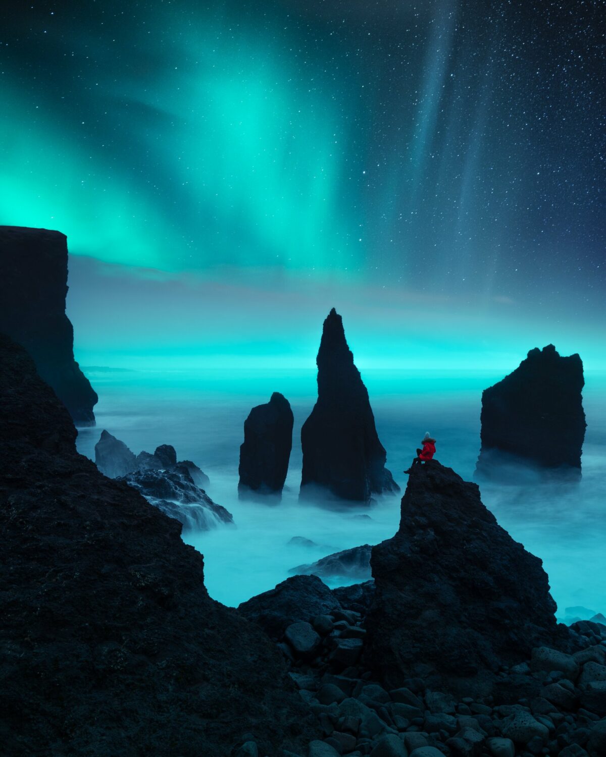 A photograph of a woman on a cliff overlooking the sea at night as blue-green aurora borealis dance above her.