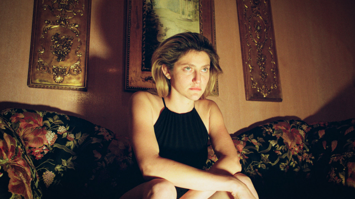 A woman wearing a black dress sits on a colorful sofa in low lamp lighting with an ominous expression on her face.