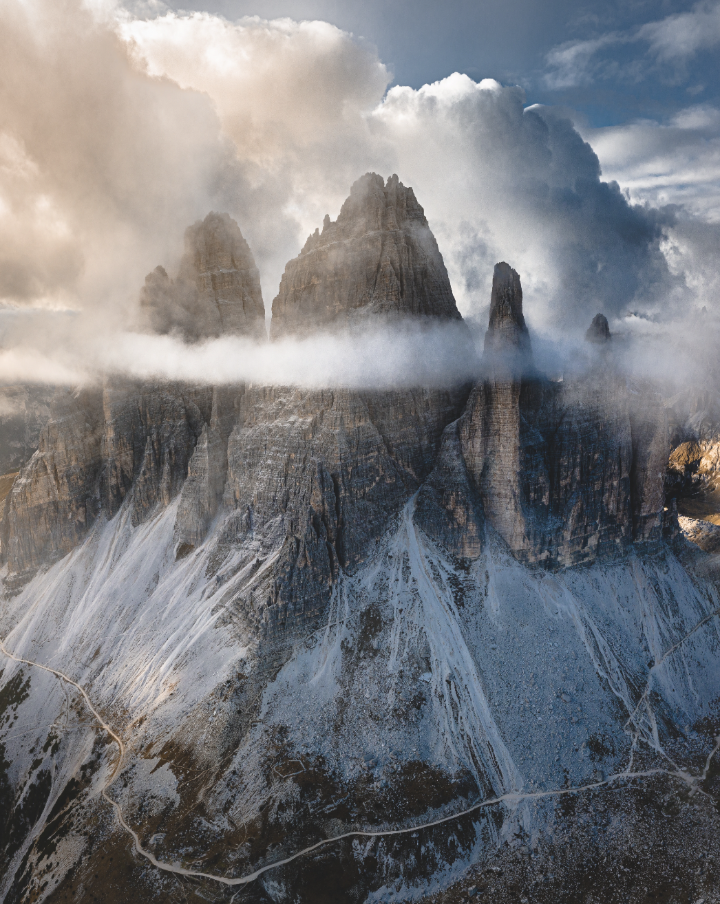 A drone photograph of the Dolomites in Italy with clouds hovering behind and above the snowy peaks.