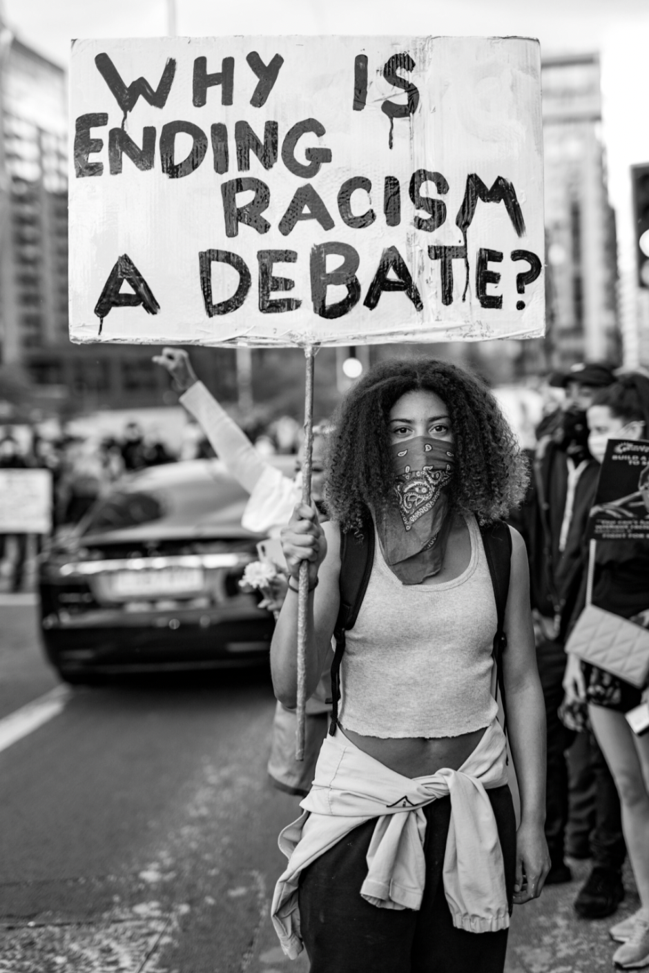 A black and white photograph of a woman wearing a bandana over her face holding a sign that says 