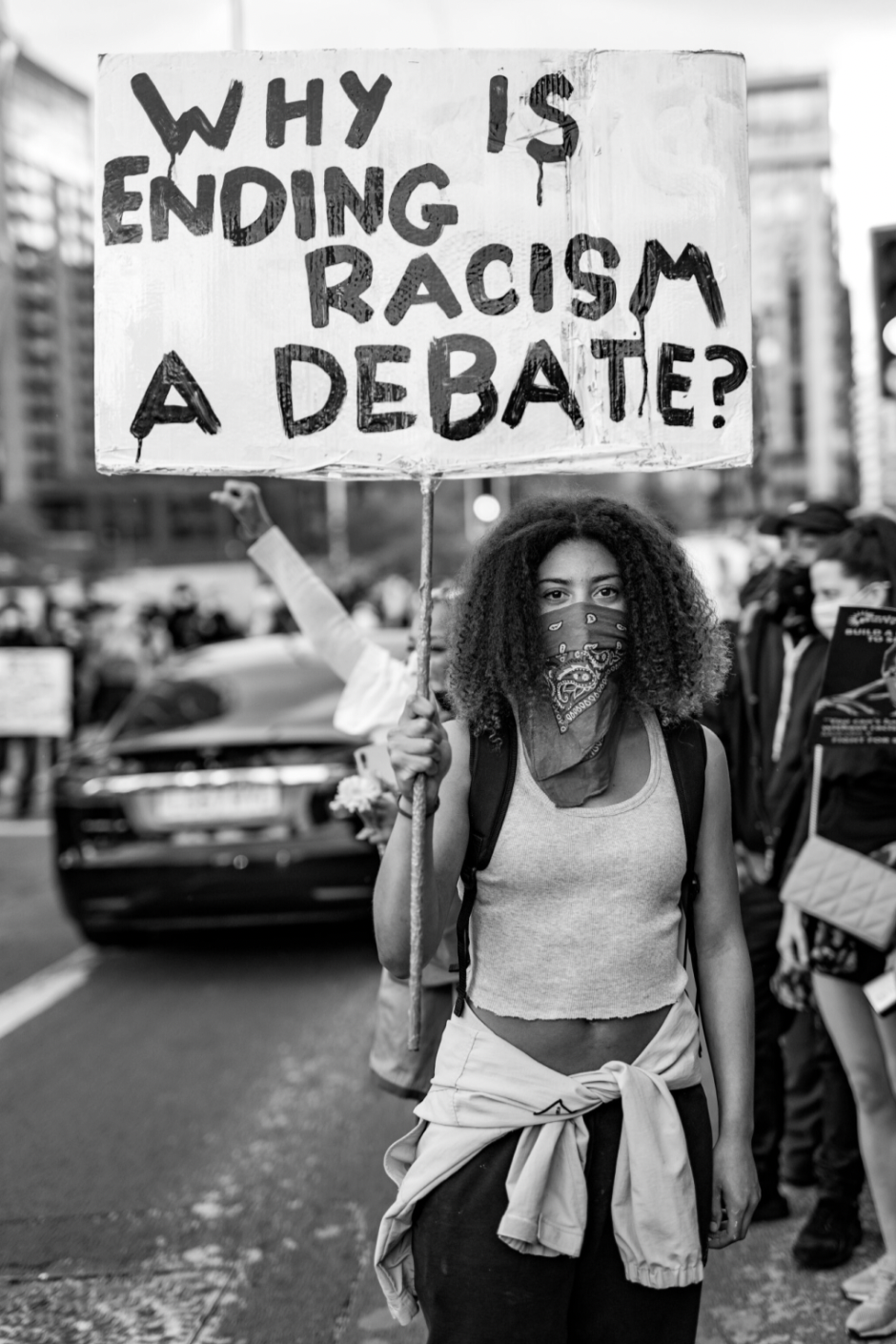 A Black And White Photograph Of A Woman Wearing A Bandana Over Her Face Holding A Sign That Says &Quot;Why Is Ending Racism A Debate?&Quot;