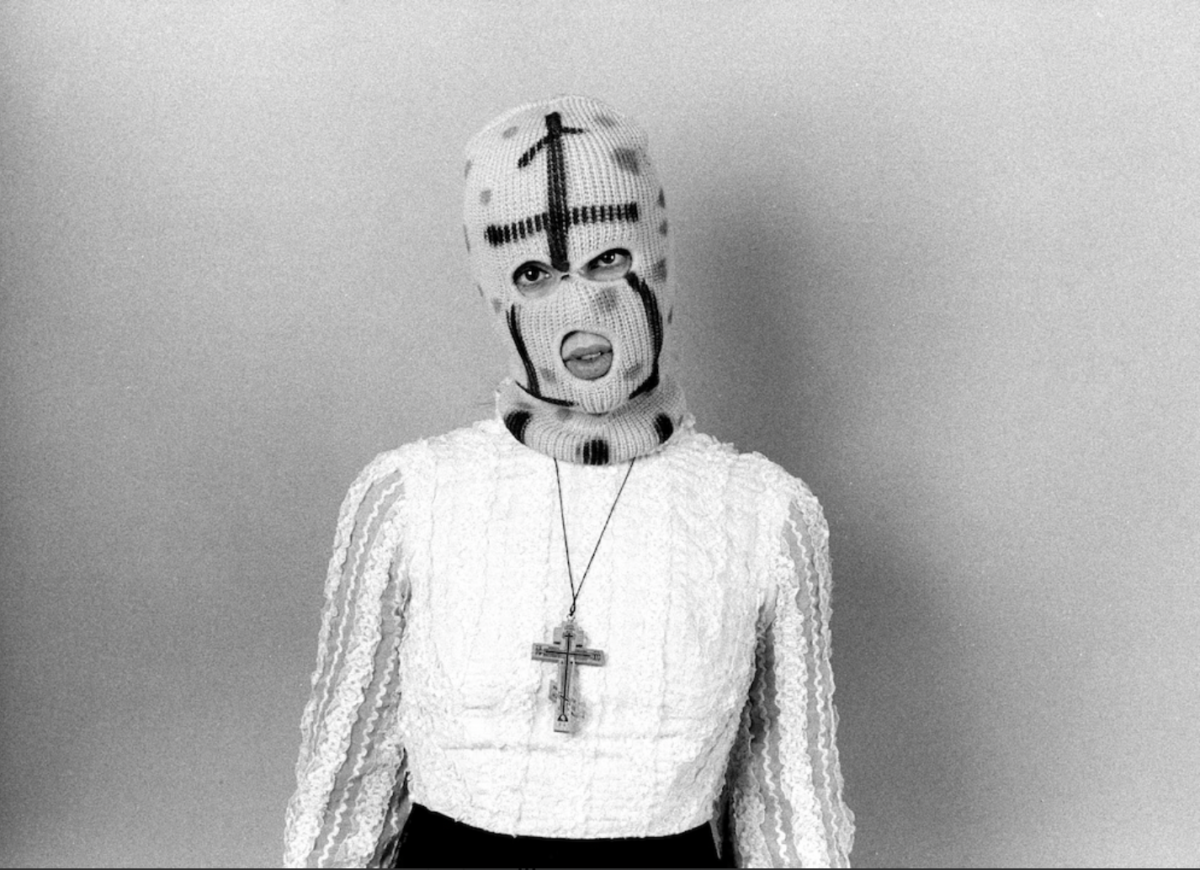 A black and white photograph of a woman wearing a white sweater and a necklace in the shape of a cross while wearing a ski mask on her head.