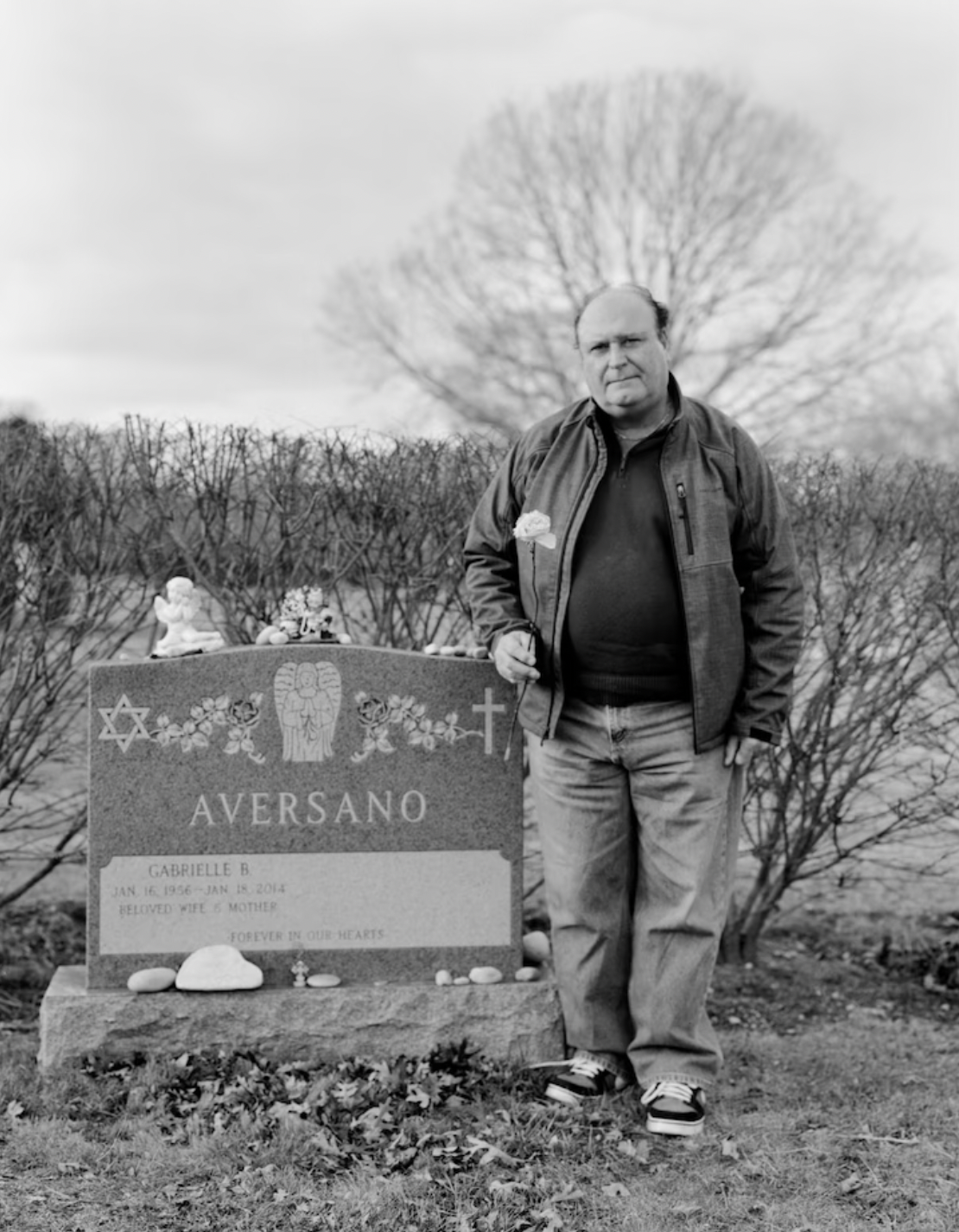 A black and white photograph of a man holding a flower standing next to the grave of his wife. There is an open space on the headstone where his name will be written when he died.