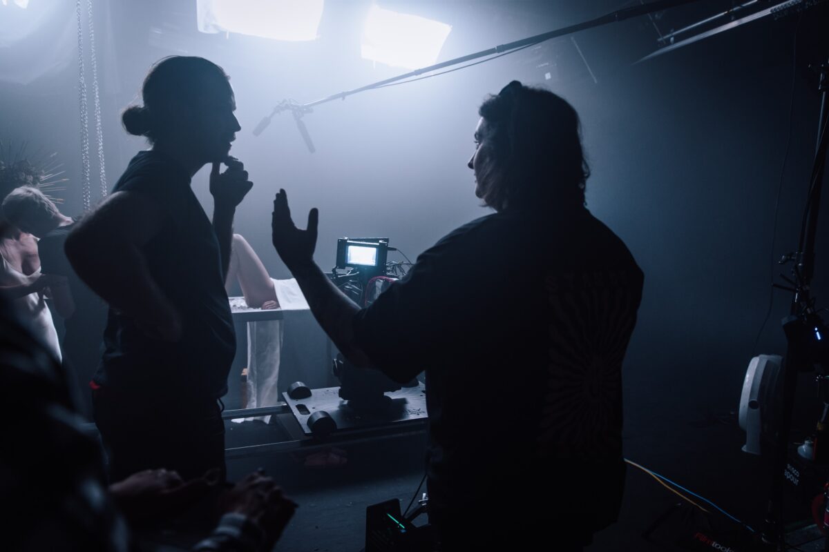 A scene from the set of a movie, strong white lights in the foreground and the silhouette of of woman and a man as they gesture and talk with one another.