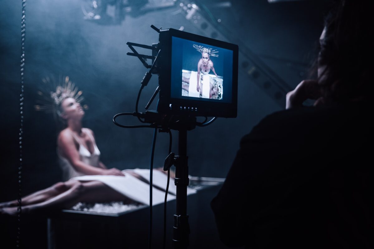A dark film set, a woman is behind a monitor watching a scene in the foreground which shows a woman wearing a white gown and a strange crown while watching over a body on a table.