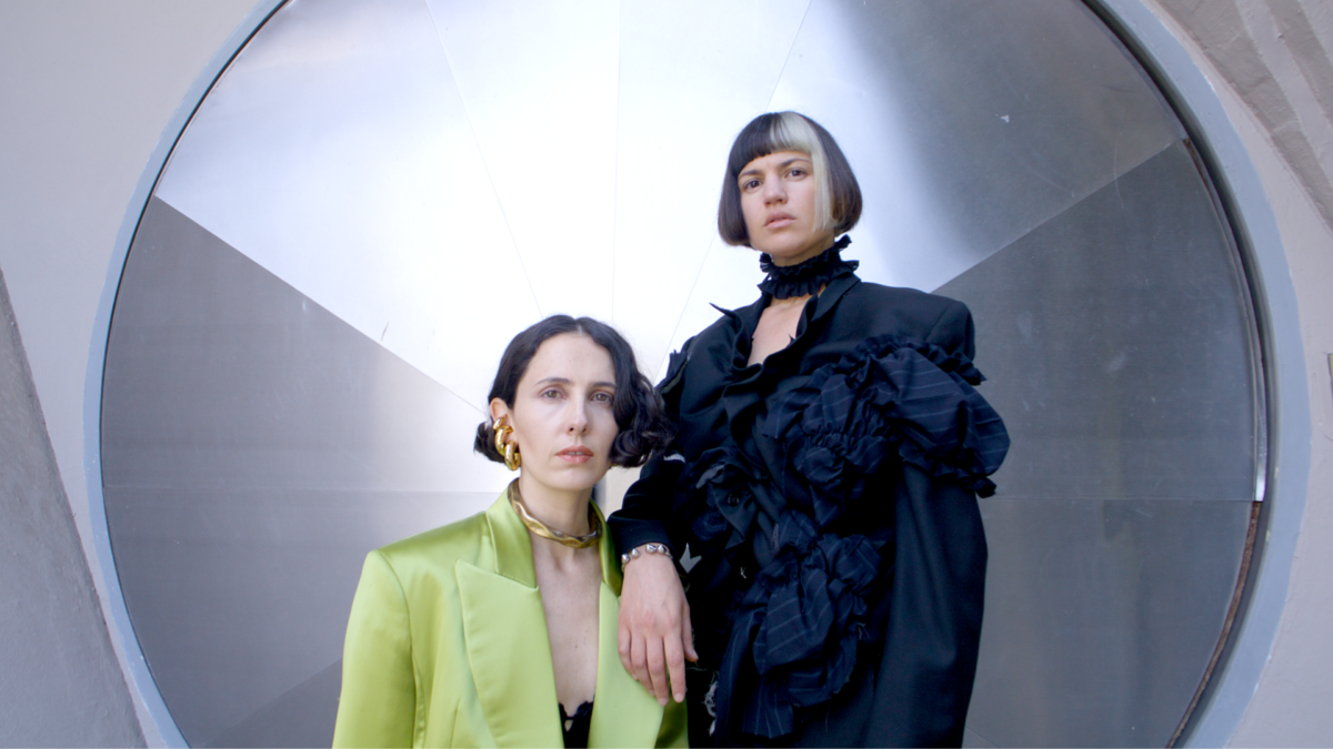 Two women, one sitting, one standing, pose for a photo in front of a large circular mirror. The woman on the left is wearing a light green colored jacket and the woman on the right is wearing a dark grey and black gown.