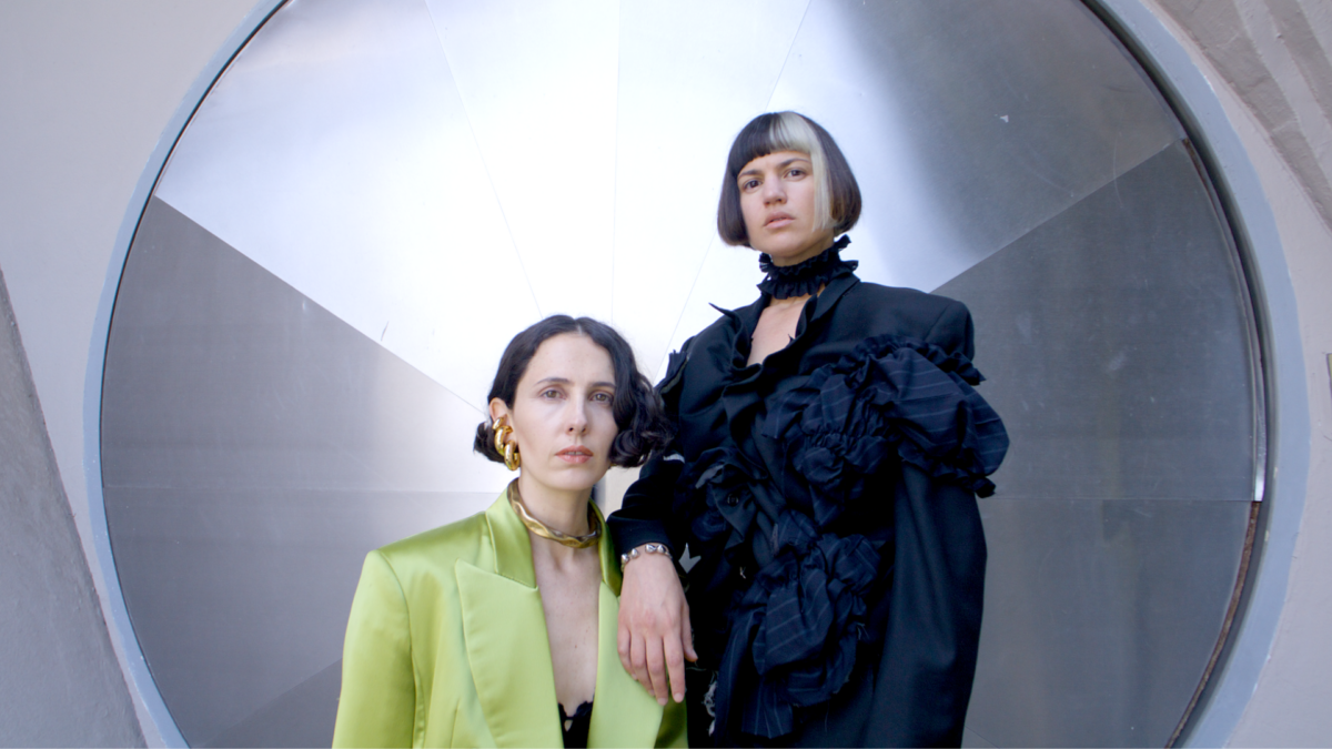 Two women, one sitting, one standing, pose for a photo in front of a large round mirror.  The woman on the left wears a light green jacket and the woman on the right wears a dark gray and black dress.