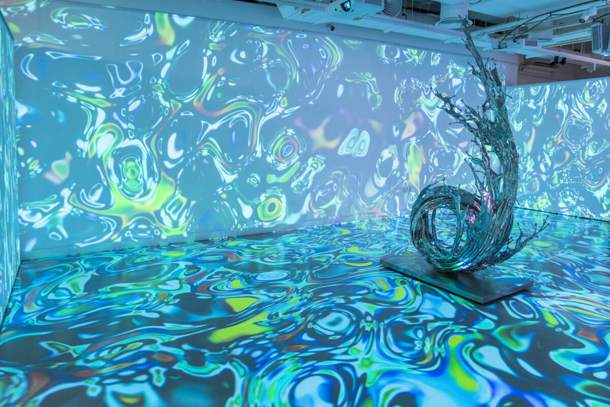 An abstract projection of blue-green shapes and colors on the wall and floor of an exhibition space featuring a wave-shaped sculpture in the middle of the room.