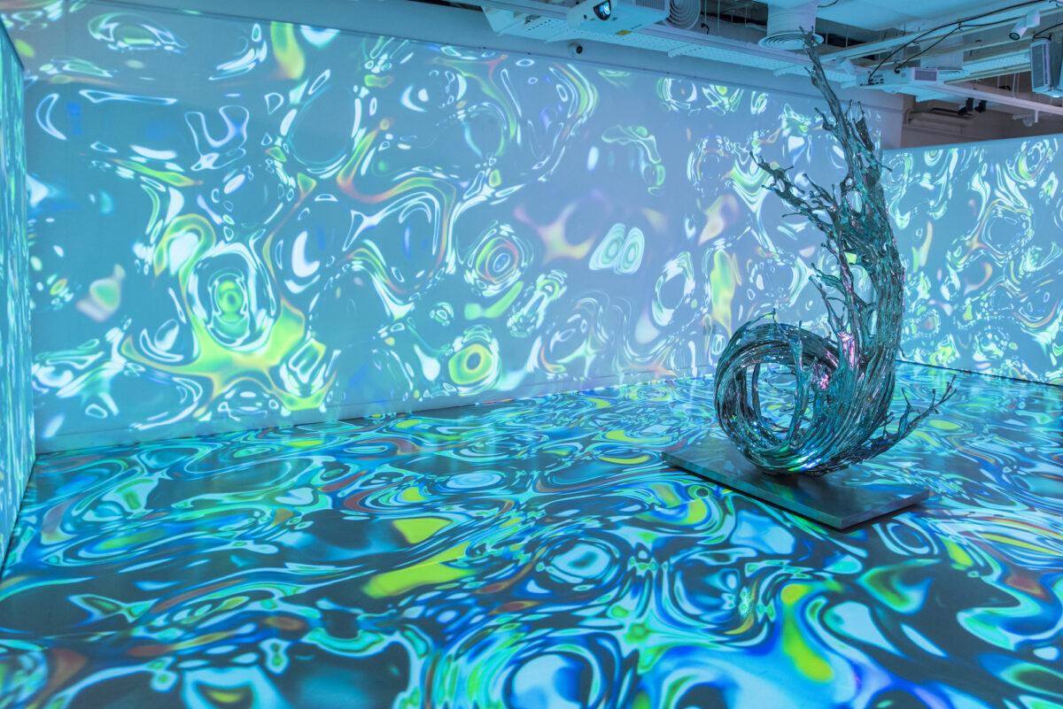 An abstract projection of blue-green shapes and colors on the wall and floor of an exhibition space with a wave-shaped sculpture in the center of the room.