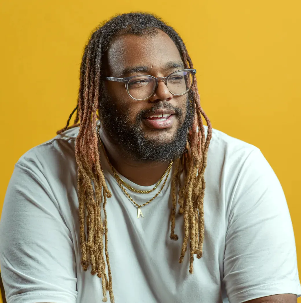 A man with dreads and glasses wearing a white t-shirt against a yellow orange background,