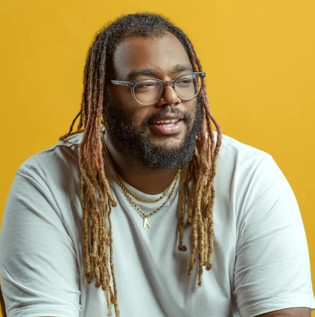 Een man met dreadlocks en een bril die een wit t-shirt draagt ​​tegen een geeloranje achtergrond,